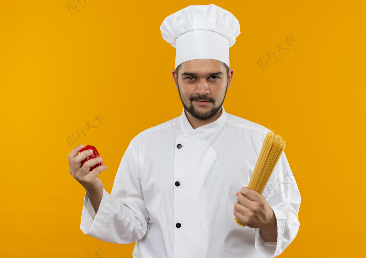 男性自信的年轻男性厨师身着厨师制服 手拿胡椒粉和意大利面 隔离在橙色的墙上制服持有意大利面