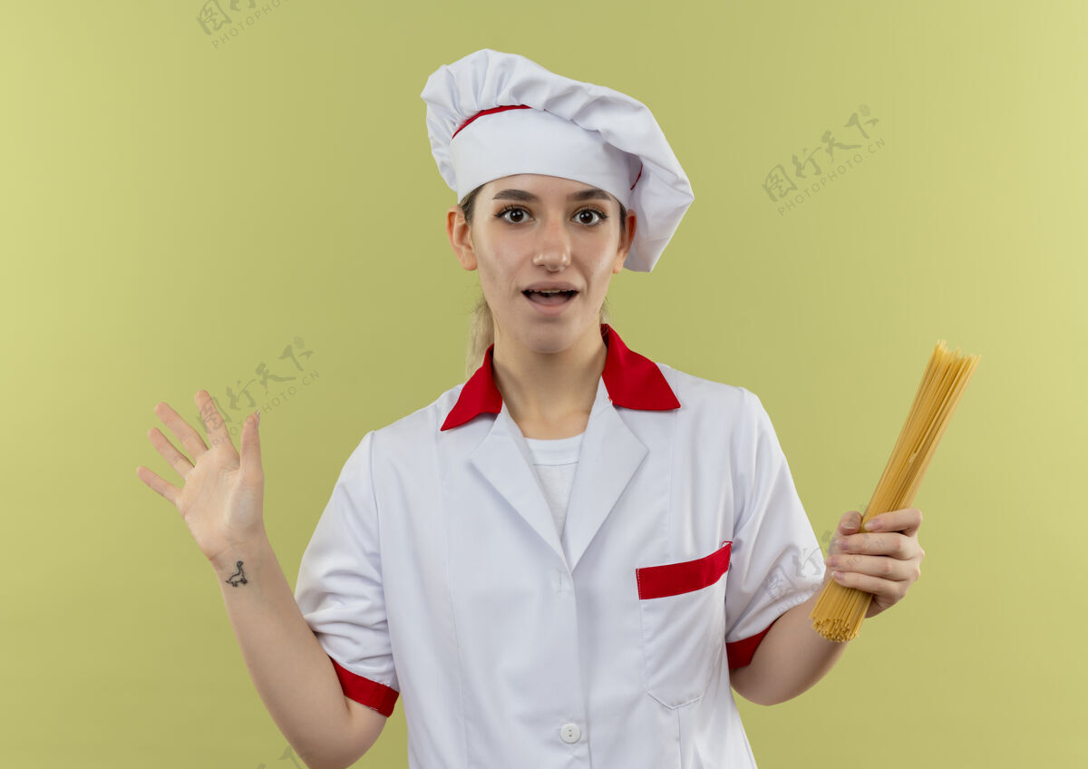 空年轻漂亮的厨师穿着厨师制服 手拿意大利面 空荡荡的手被隔离在绿色的墙上持有显示意大利面