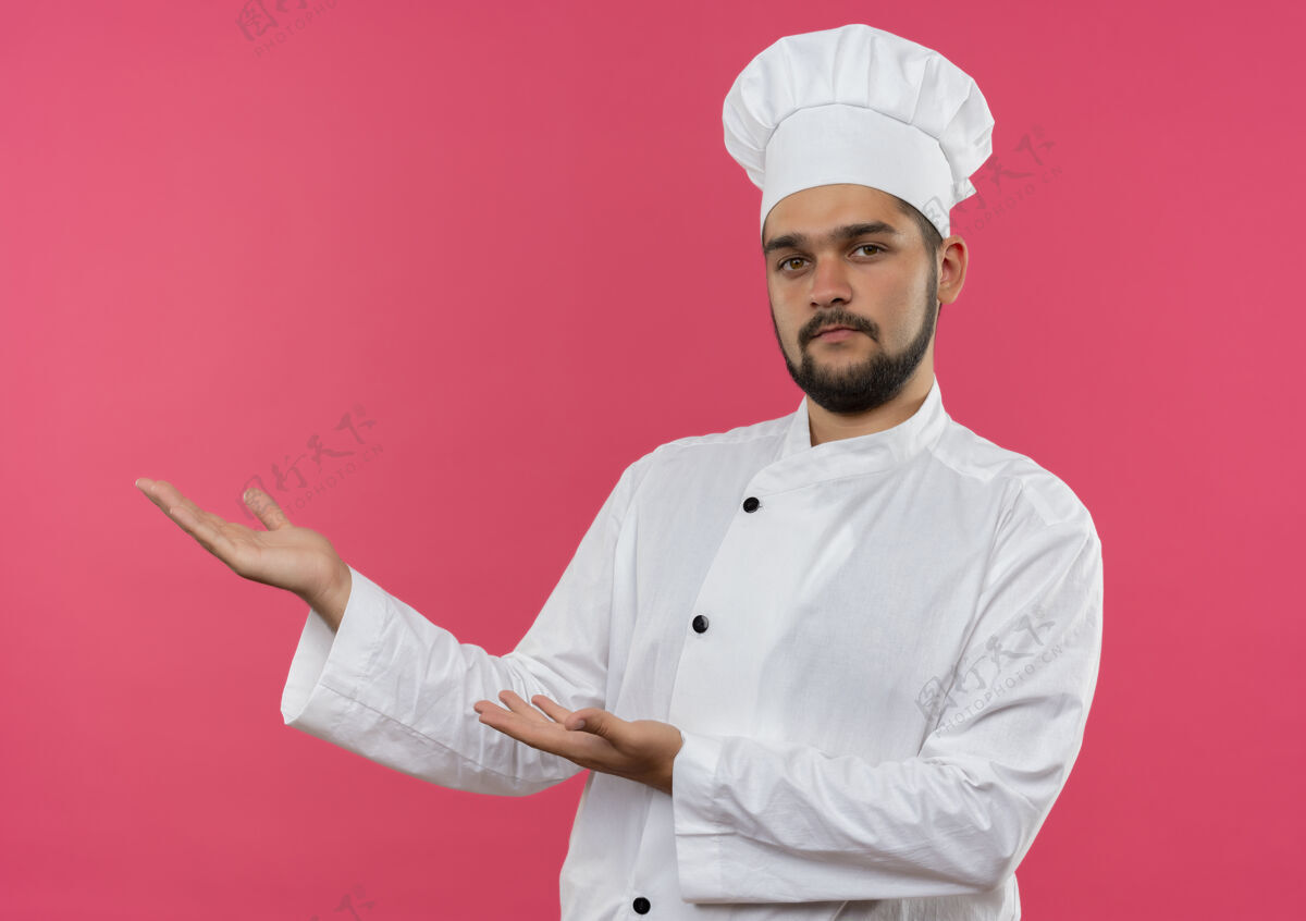 烹饪自信的年轻男性厨师 身着厨师制服 双手指向隔离在粉红色墙上的一侧指点制服壁板