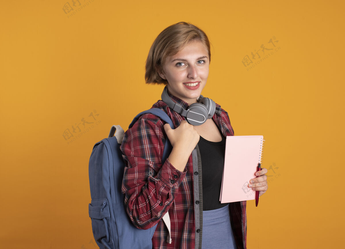 空间微笑的年轻斯拉夫女学生戴着耳机 背着背包 拿着笔记本和笔 看着相机钢笔穿微笑