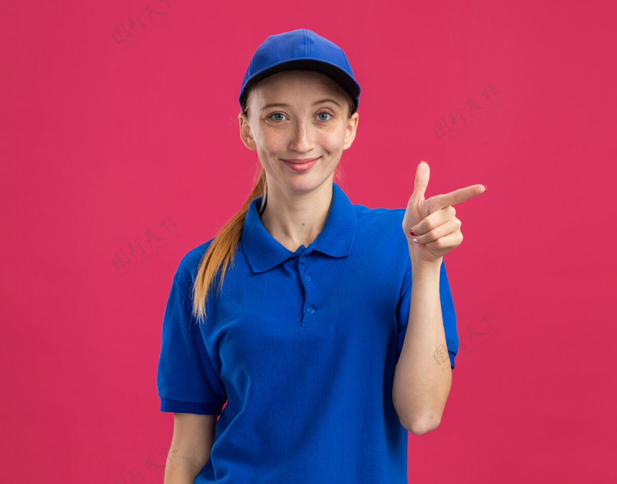微笑身穿蓝色制服 头戴鸭舌帽的年轻送货员友好地用食指指着站在粉色墙上的一侧制服指点朋友