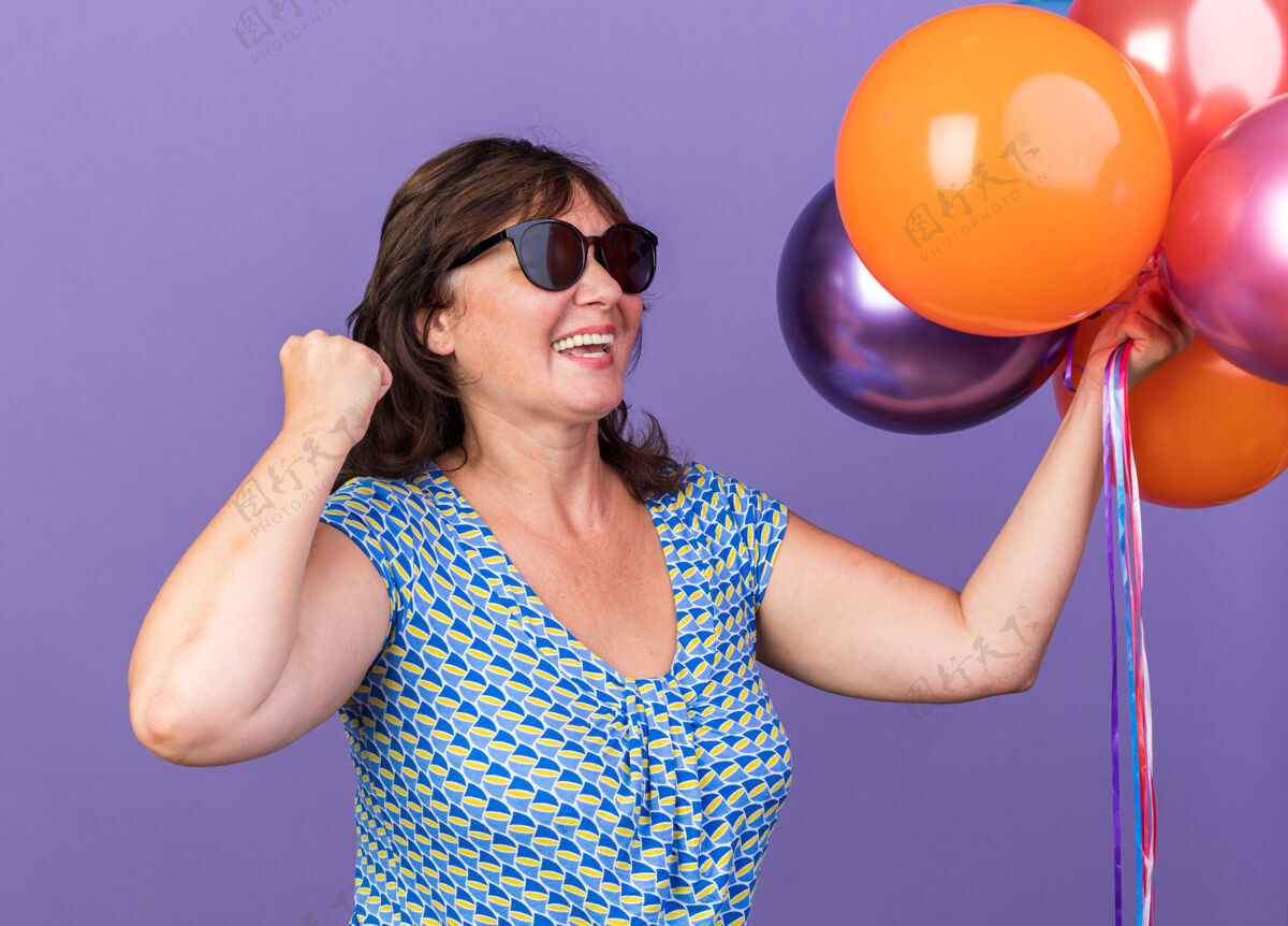 立场快乐和兴奋的中年妇女手持一束五颜六色的气球握紧拳头庆祝生日聚会站在紫色的墙上握紧庆祝生日
