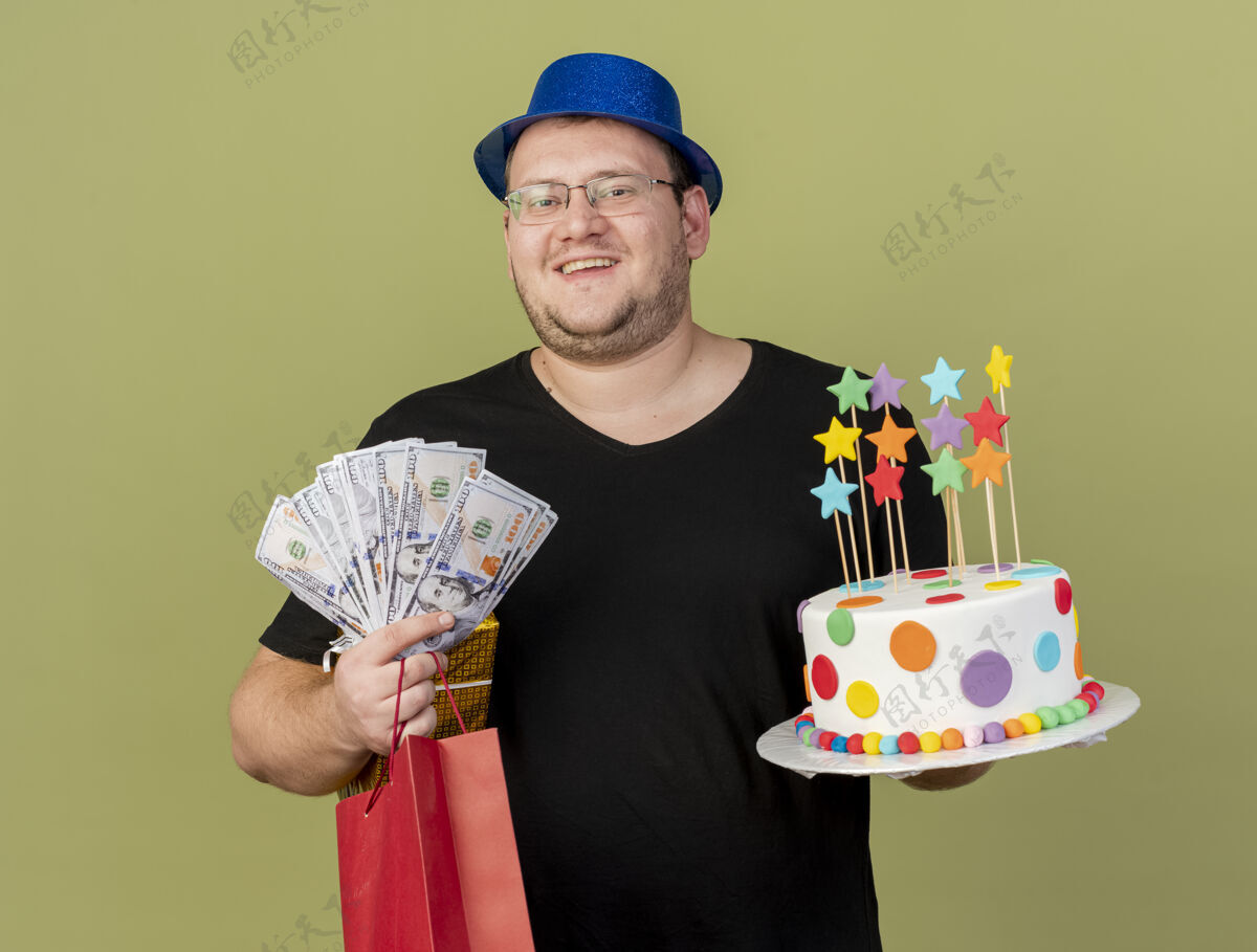 包微笑的成年斯拉夫人戴着眼镜 戴着蓝色派对帽 手里拿着钱礼盒 纸购物袋和生日蛋糕橄榄持有成人