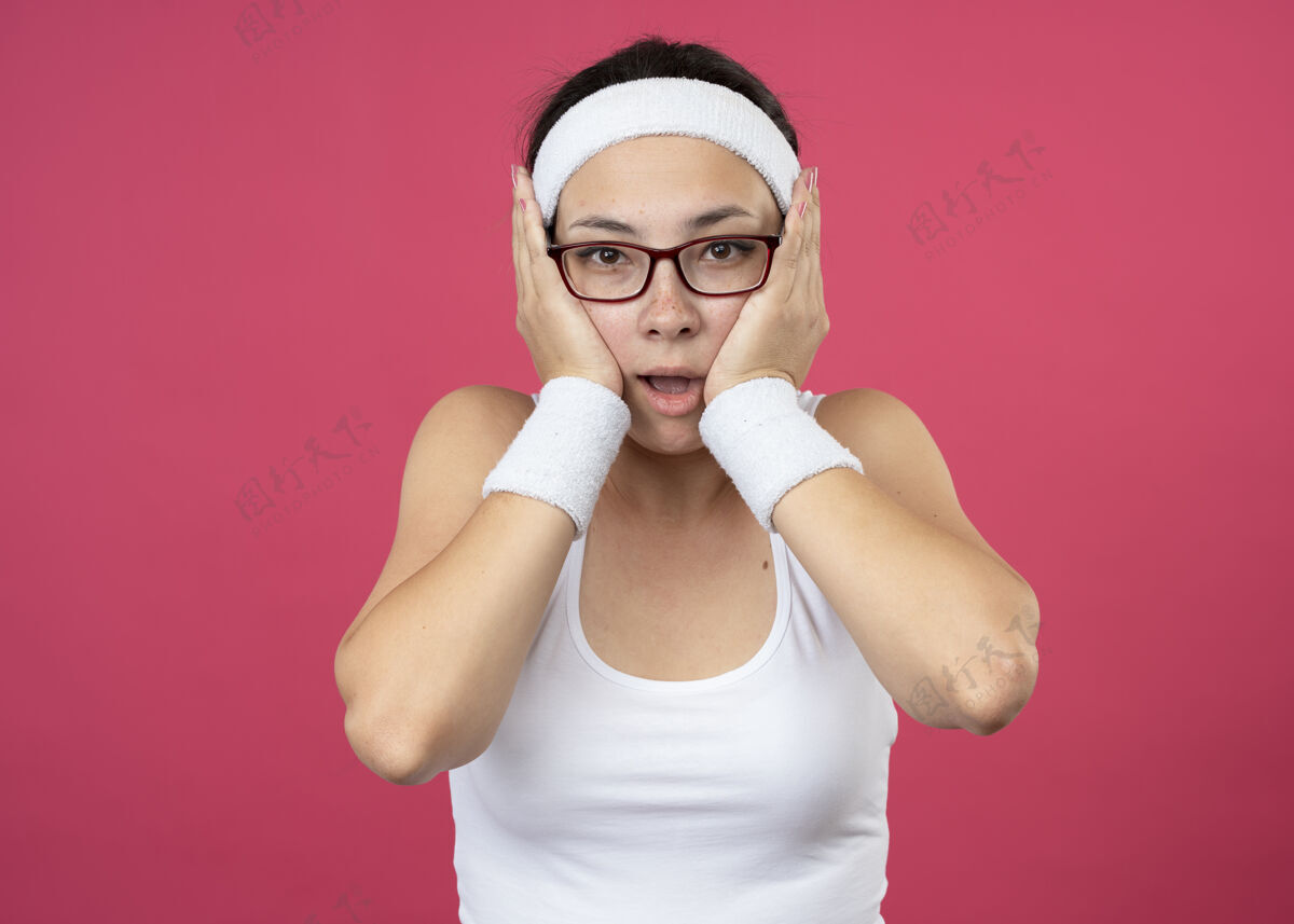 脸兴奋的年轻运动女孩戴着眼镜戴着头带和腕带把手放在脸上佩戴腕带兴奋