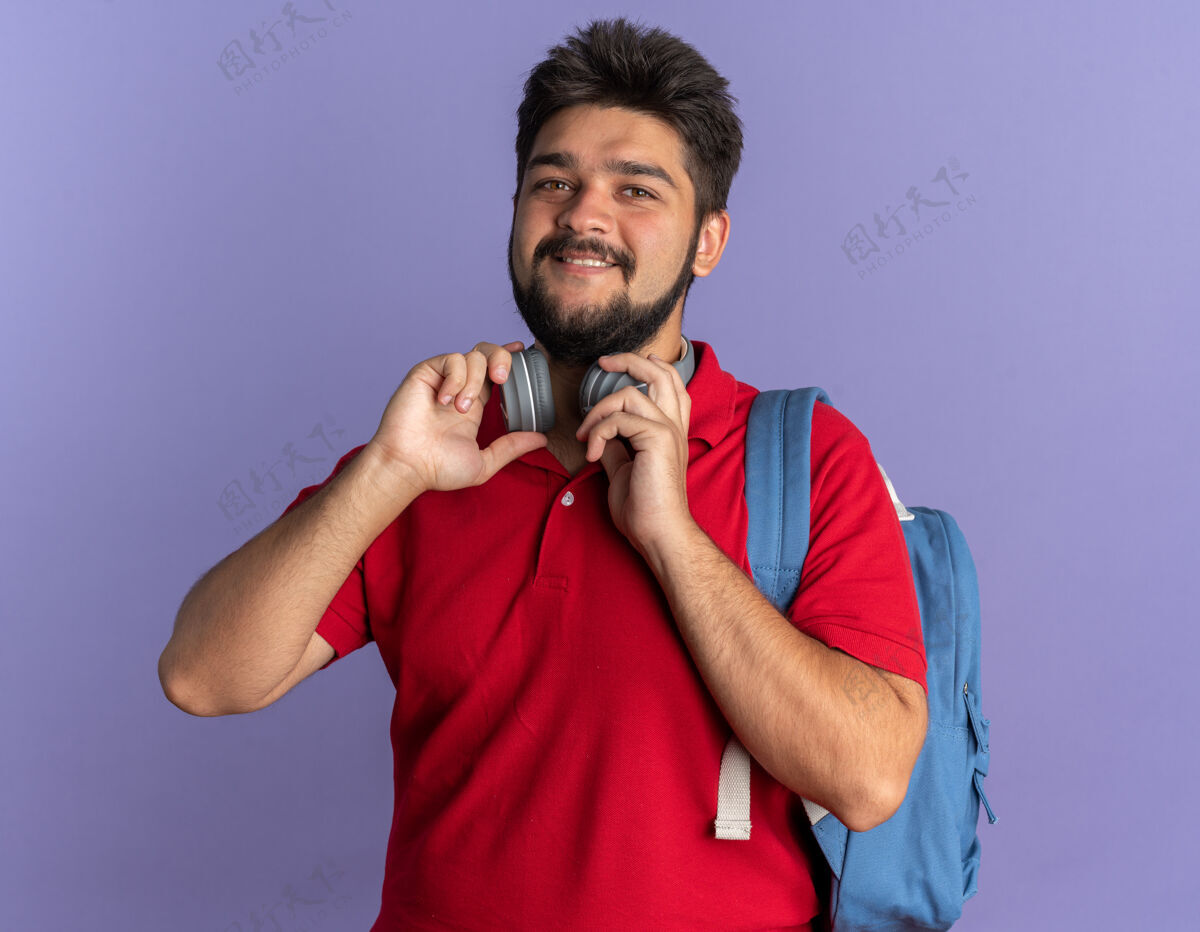 学生年轻的留着胡子的学生 穿着红色马球衫 背着背包 戴着耳机 面带微笑 站在那里快乐而积极胡须小伙子欢呼