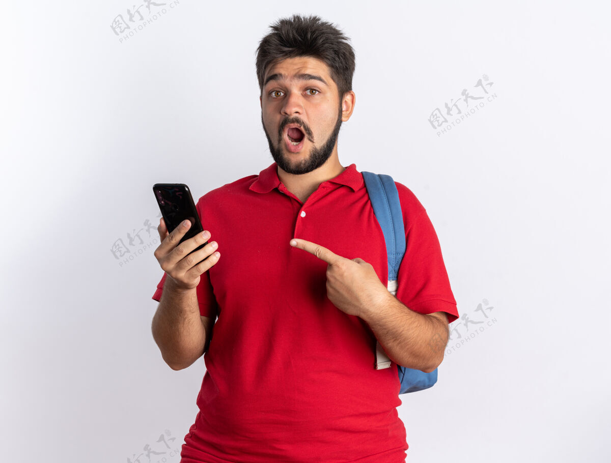 指着一个年轻的留着胡子的学生 穿着红色马球衫 背着背包 拿着智能手机 用食指指着它 站在那里显得很惊讶站着背包胡子