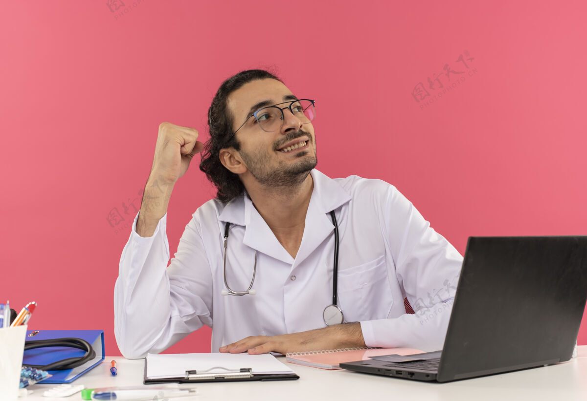 空间面带微笑的年轻男医生戴着医用眼镜 穿着医用长袍 手持听诊器坐在办公桌旁医生微笑工作