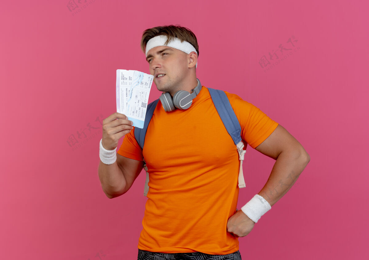 包体贴的年轻帅气的运动型男人 戴着头带和腕带 背着一个包 脖子上戴着耳机 手放在腰上 手里拿着机票 一边看一边粉色飞机腕带