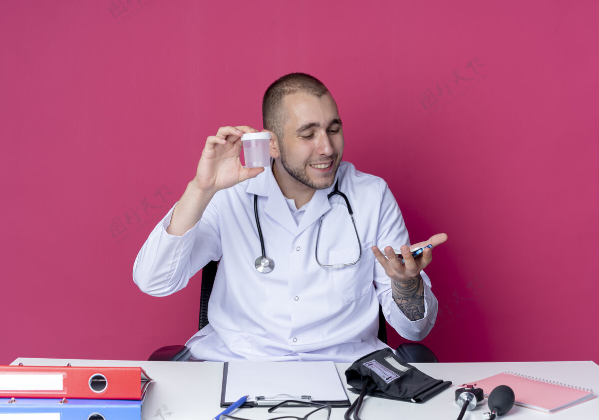 药片微笑的年轻男医生穿着医用长袍和听诊器坐在办公桌旁包工作男性