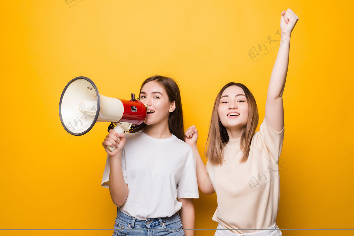 一起两个恼怒的年轻女子女孩朋友在黄色墙壁上用扩音器尖叫人们的生活方式概念模拟复制空间人朋友脸