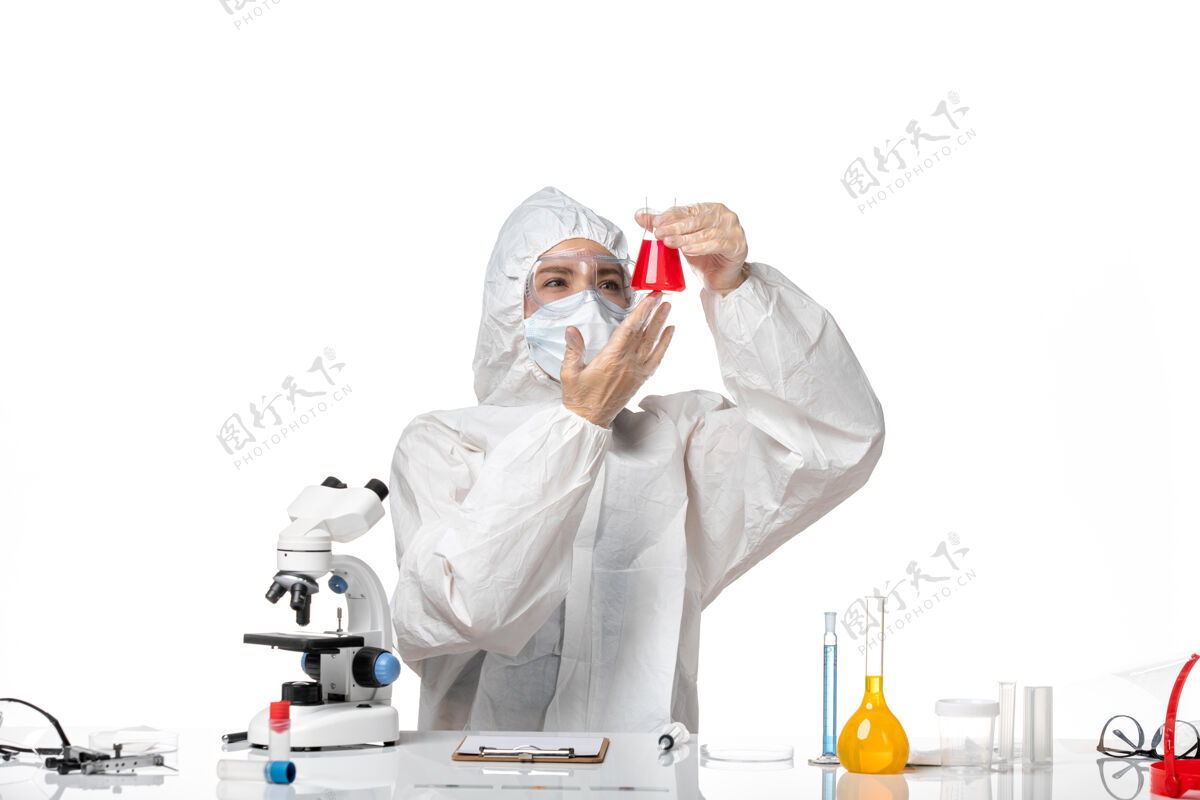 专业正面图年轻女医生穿着白色防护服带口罩由于柯维德手持红色溶液在白色办公桌上病毒大流行飞溅柯维德-面罩药病毒