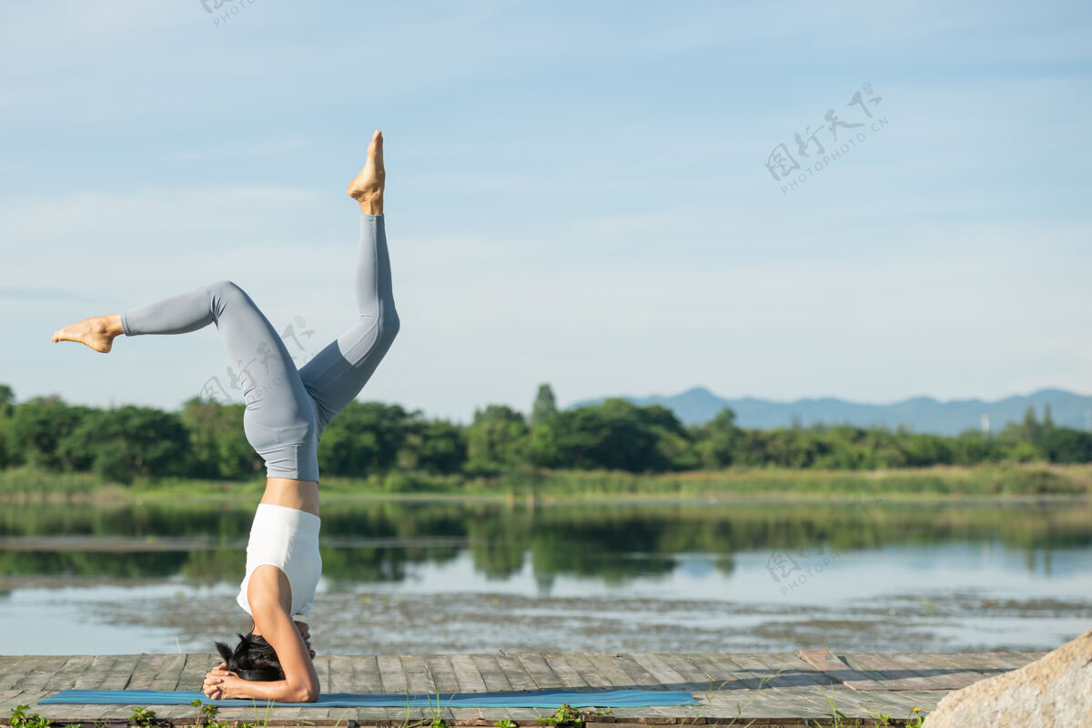 阳光坐在瑜伽垫上在公园里放松的女人年轻的运动型亚洲女人练习瑜伽 做倒立运动 锻炼身体 穿着运动服 裤子和上衣河流自然健身