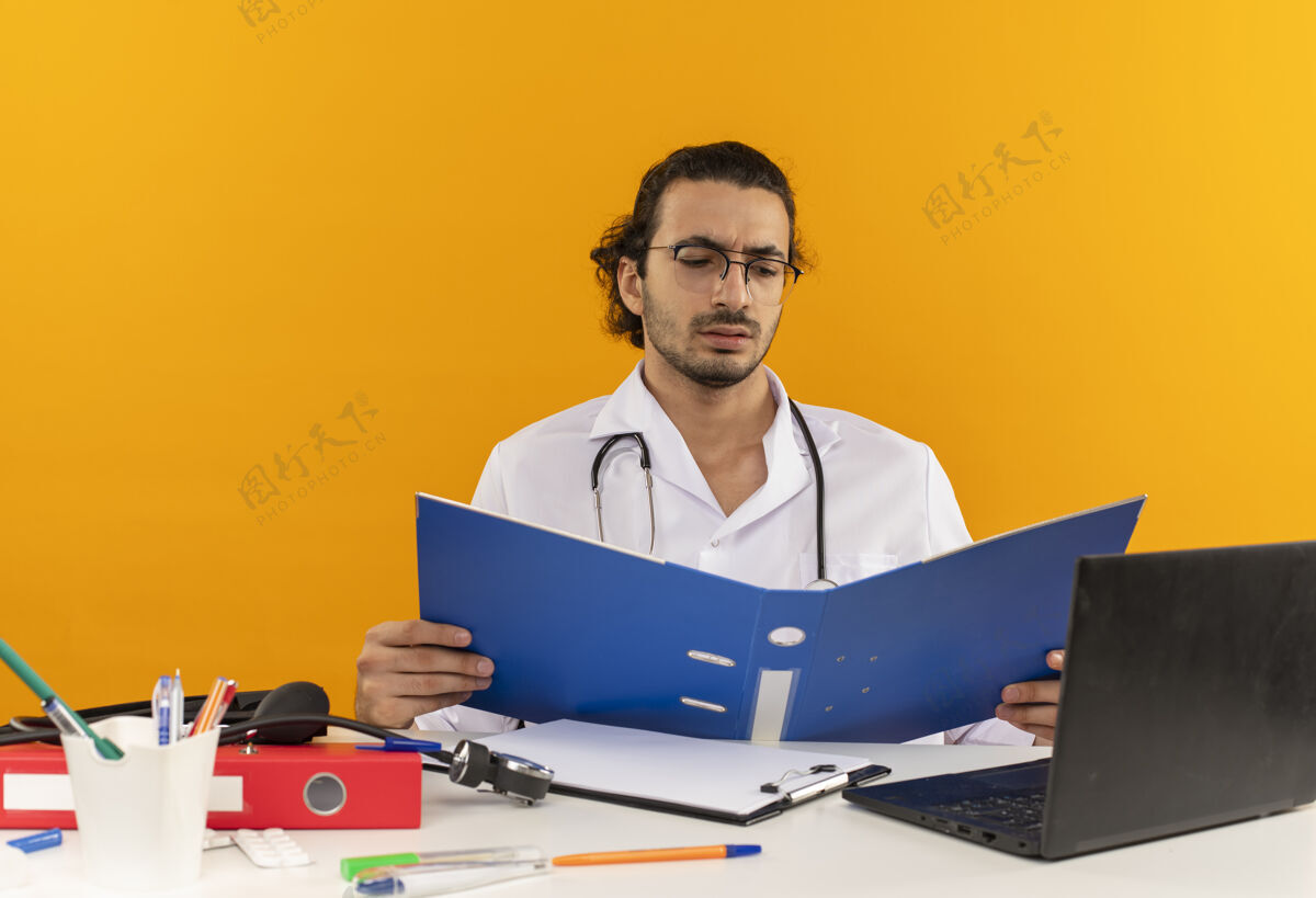 听诊器年轻的男医生戴着医用眼镜 穿着医用长袍 听诊器坐在桌边办公桌医疗工作