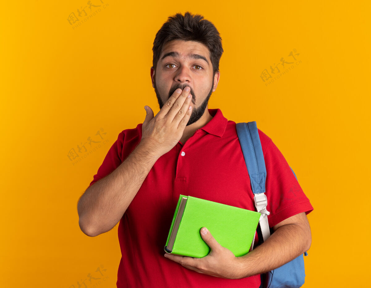 背包年轻的留着胡子的学生 穿着红色马球衫 背着背包 拿着笔记本 看起来很震惊 手站着捂着嘴学生胡子笔记本