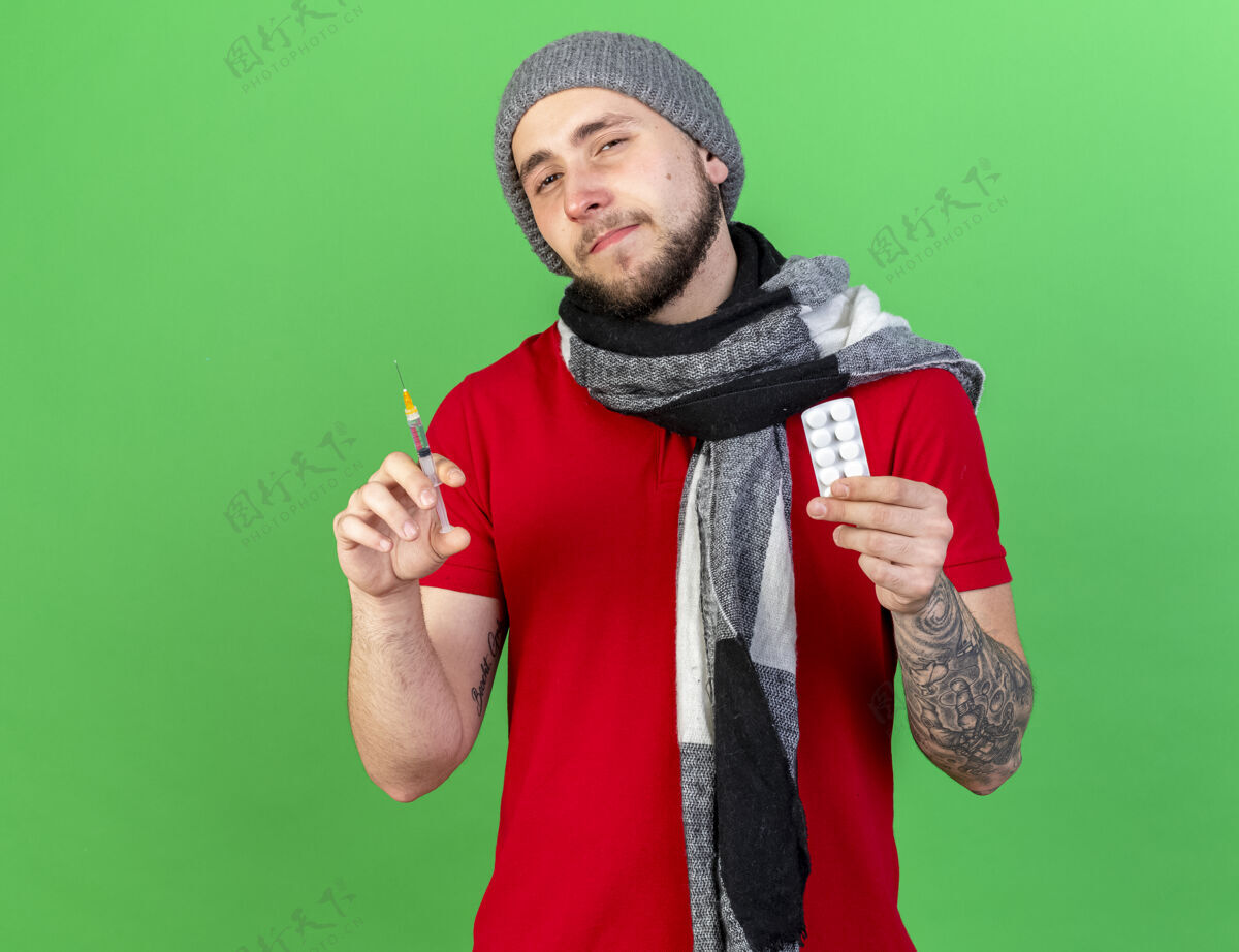 穿自信的年轻白种人 戴着冬天的帽子 戴着围巾 手里拿着注射器和一包药片 站在绿色的草地上男人年轻医学