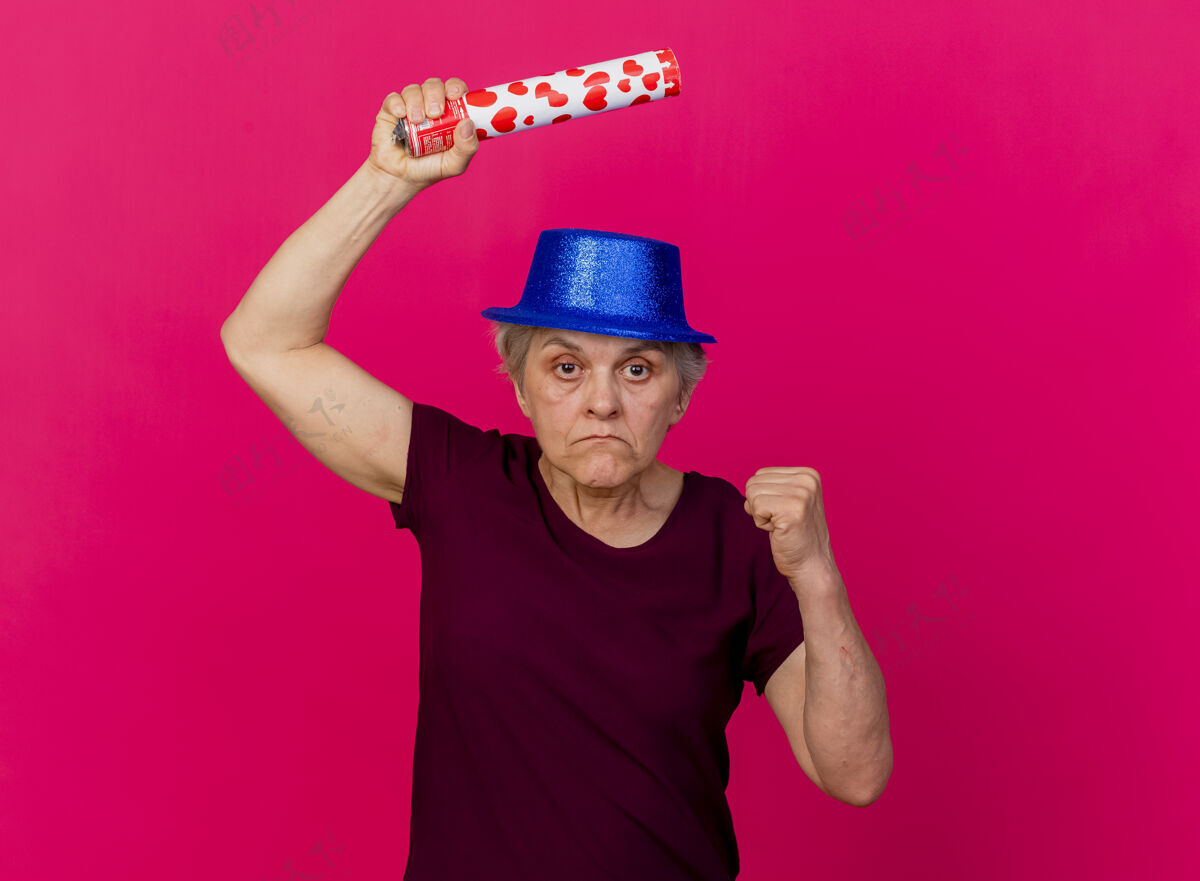 帽子戴着派对帽的自信的老妇人手持五彩纸屑大炮 把拳头放在粉色的脸上派对拳头五彩纸屑