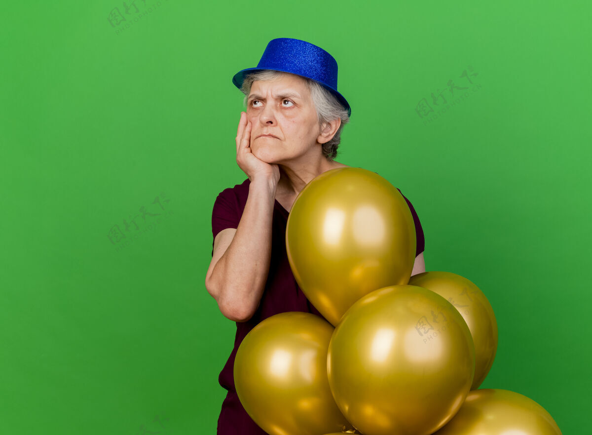 站一位戴着派对帽的老妇人把手放在下巴上 手里拿着氦气球站着 看着绿色的一面不愉快壁板氦