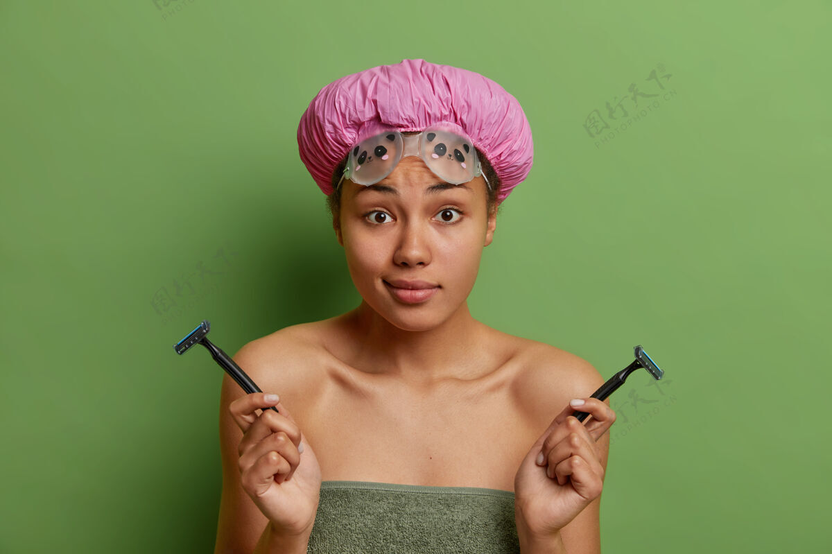 帽子迷糊的女人戴着防水帽拿着两把剃须刀去做脱毛用浴巾包着隔离在绿色的墙上剃刀人体不知道