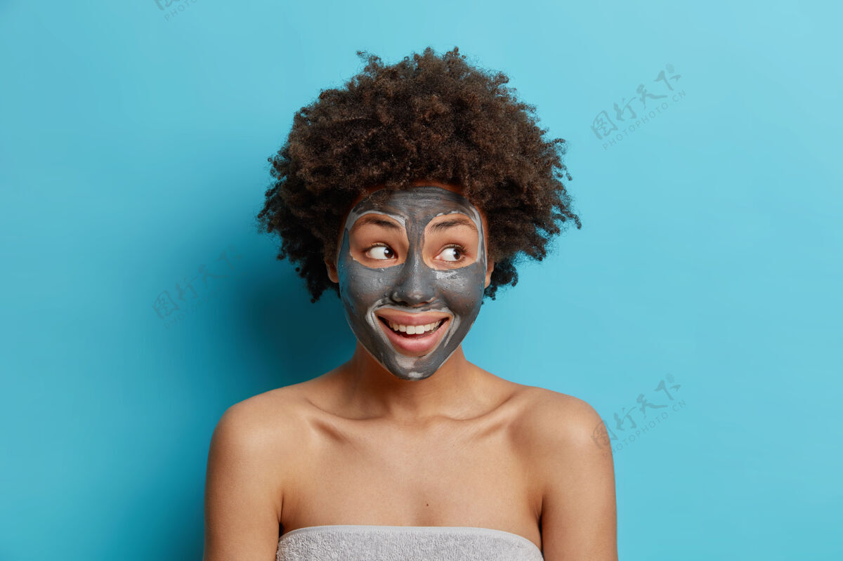 身体面部护理概念积极的卷发女人在脸上涂上粘土面膜 使皮肤恢复活力在沐浴后 用毛巾包裹在蓝色墙壁上 进行美容程序卷发包裹清洁