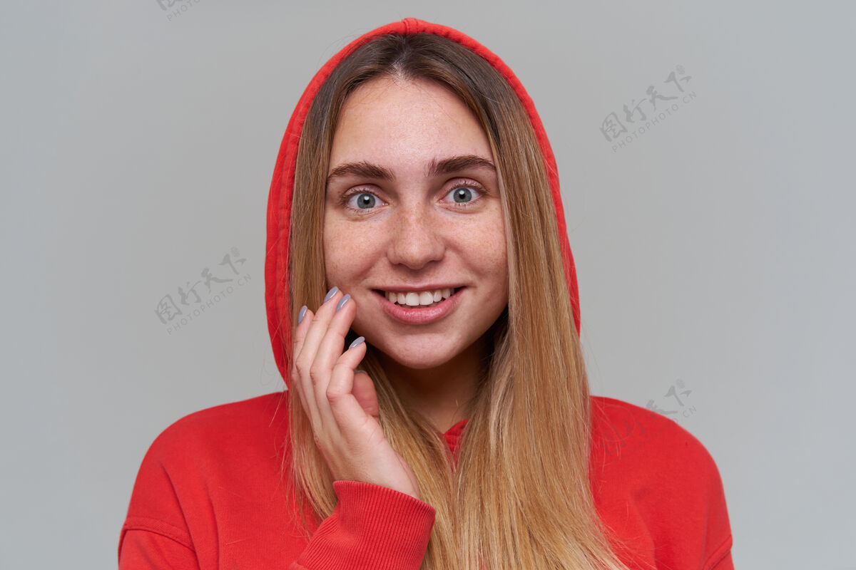 人特写肖像od快乐美丽的金发年轻女子在红色连帽衫触摸她的脸颊 在灰色的墙上看着相机休闲年轻白皙