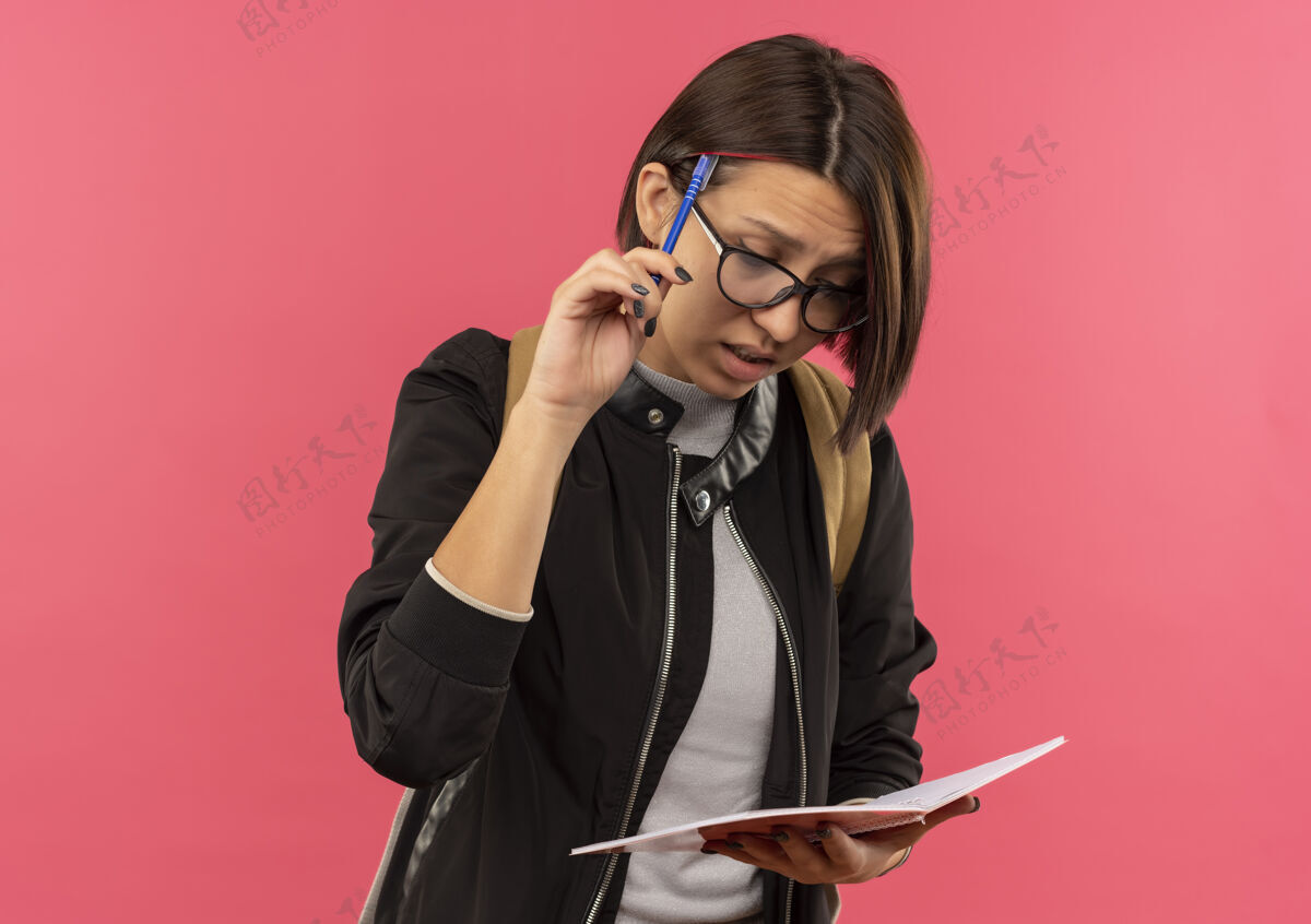 脸体贴的年轻女学生戴着眼镜 背着书包 拿着笔记本 看着笔触着头 隔离在粉红色的墙上表情握人