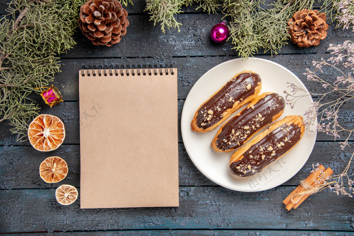 餐厅俯视图巧克力eclairs在白色椭圆形板冷杉树枝和锥圣诞玩具干花枝干桔子肉桂和一个笔记本在黑暗的木桌上盘子肉桂午餐