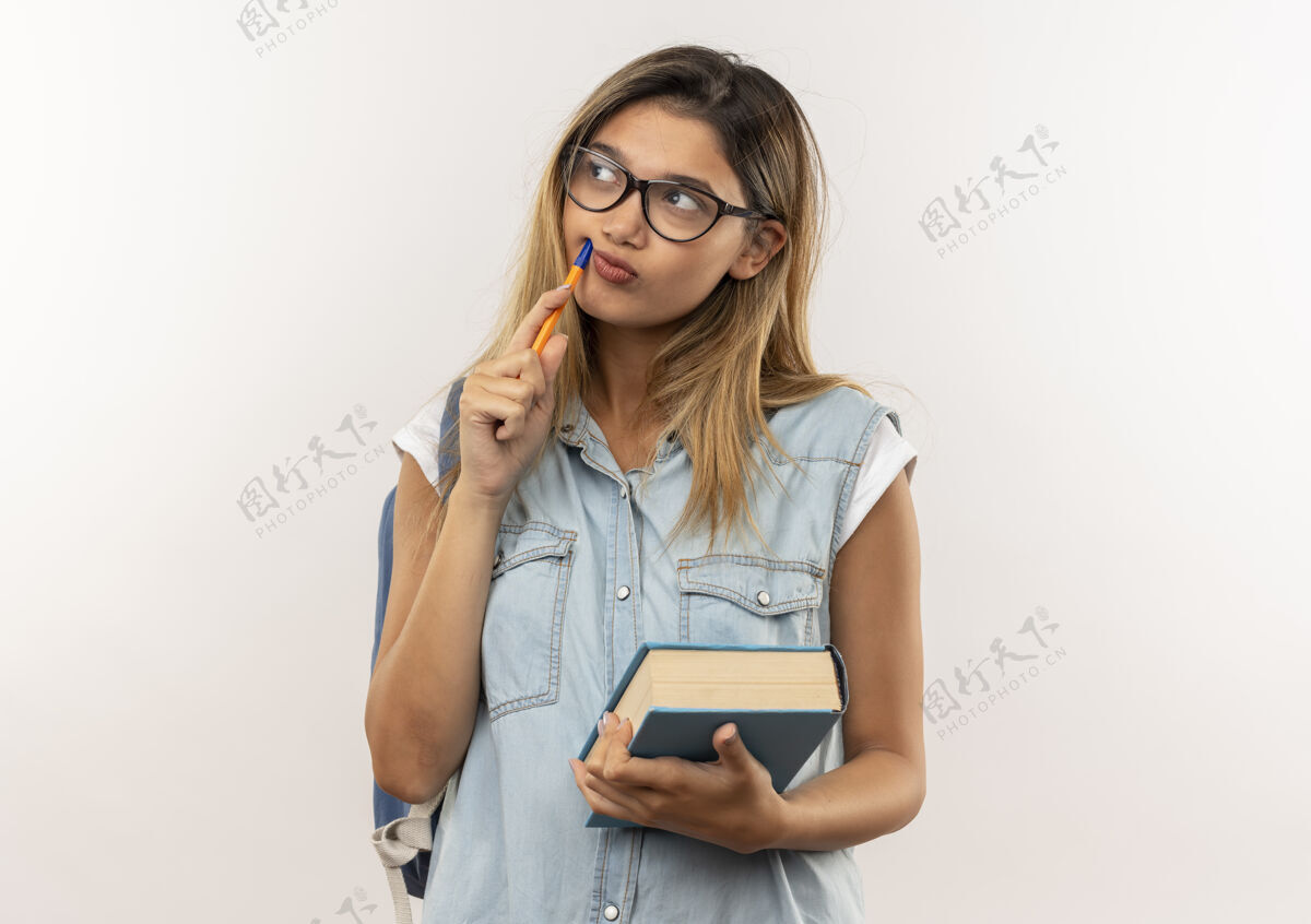 漂亮体贴的年轻漂亮的女学生戴着眼镜 背着书包 拿着书 用笔触摸着脸 看着隔离在白墙上的一面市民人思想