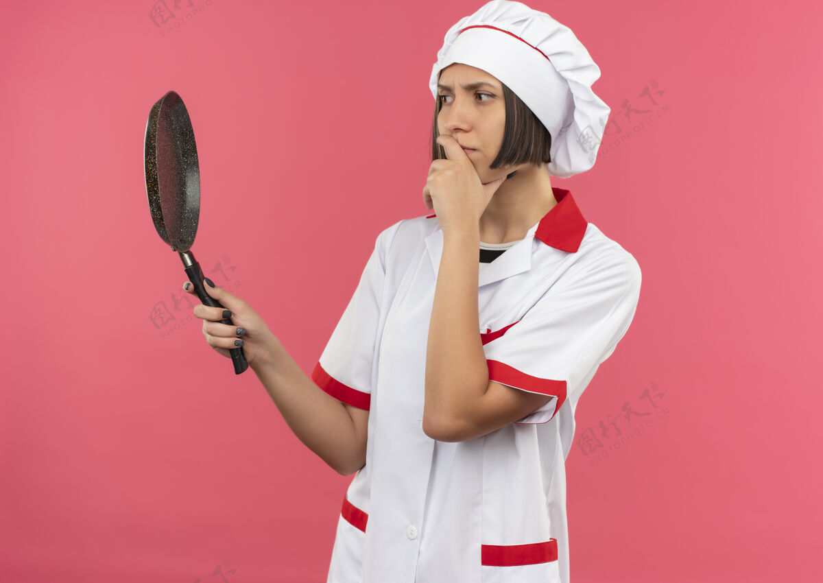 下巴体贴的年轻女厨师 穿着厨师制服 手放在下巴上 手举着手 看着粉红色墙上孤立的煎锅烹饪粉色锅