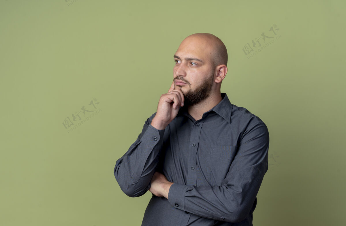 感觉体贴的年轻秃头呼叫中心男子站在橄榄绿的墙上 以封闭的姿势看着直手下巴隔离绿秃头姿势
