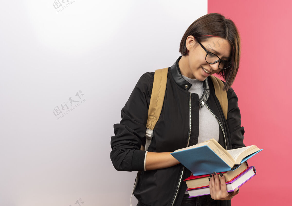 表情微笑的年轻女学生戴着眼镜 背着书包 拿着书 站在白色的墙前 孤立在粉色的墙上抱着前面人