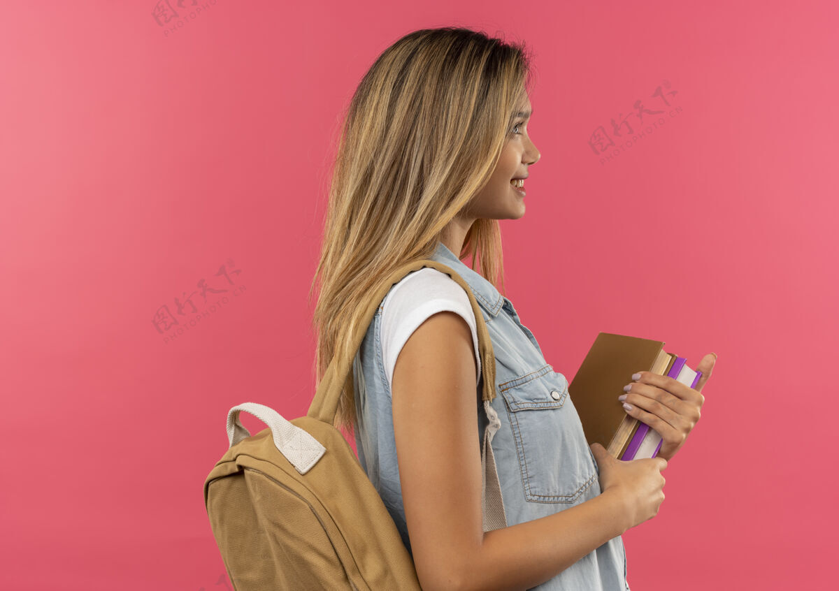 学生面带微笑的年轻漂亮的女学生 背着书包站在侧视图里 手里拿着孤立在粉红色墙上的书脸人姿势