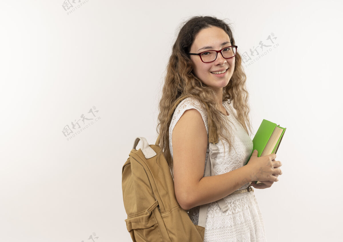 人面带微笑的年轻漂亮的女学生戴着眼镜 背着书包站在侧视图里 手里拿着孤立在白墙上的书穿人姿势