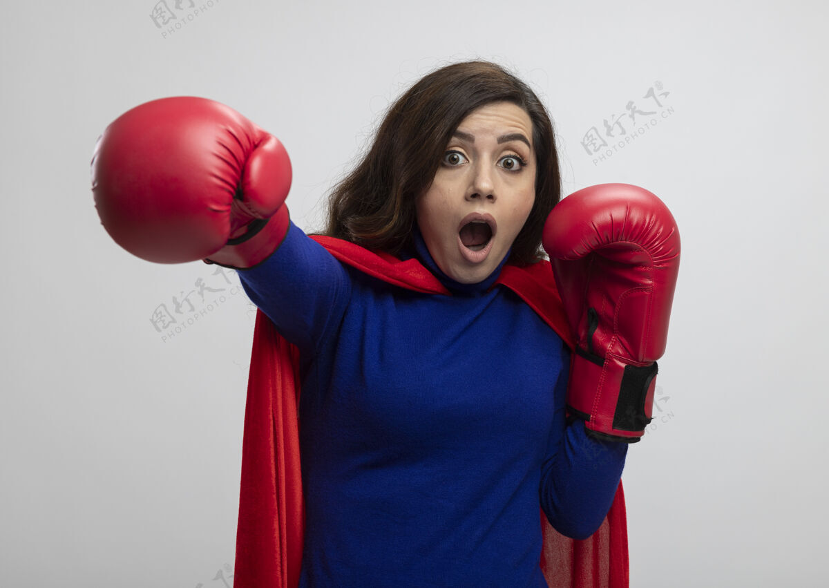 超级英雄震惊的高加索超级英雄女孩穿着红色斗篷戴拳击手套假装打拳拳击假装拳击