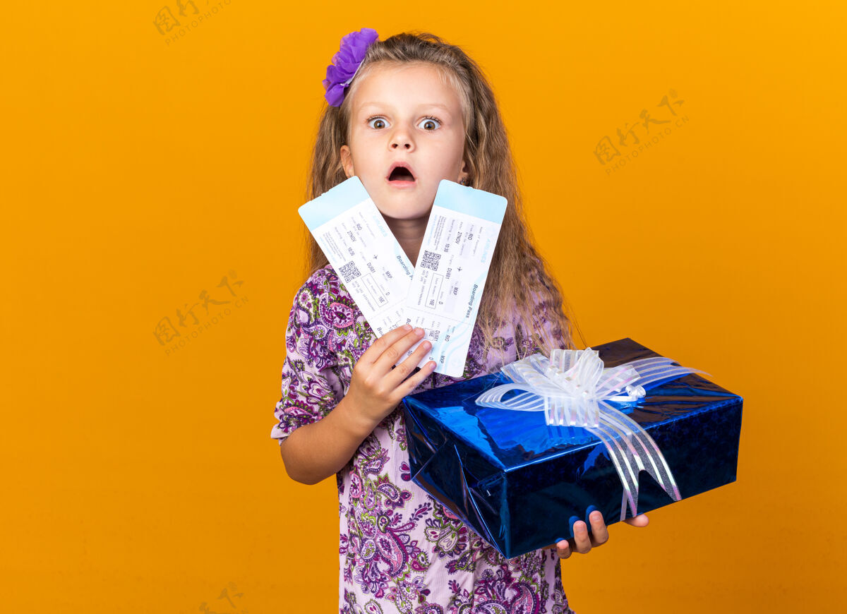 孤立震惊的金发小女孩拿着礼盒和机票隔离在橙色墙上的复制空间飞机机票小