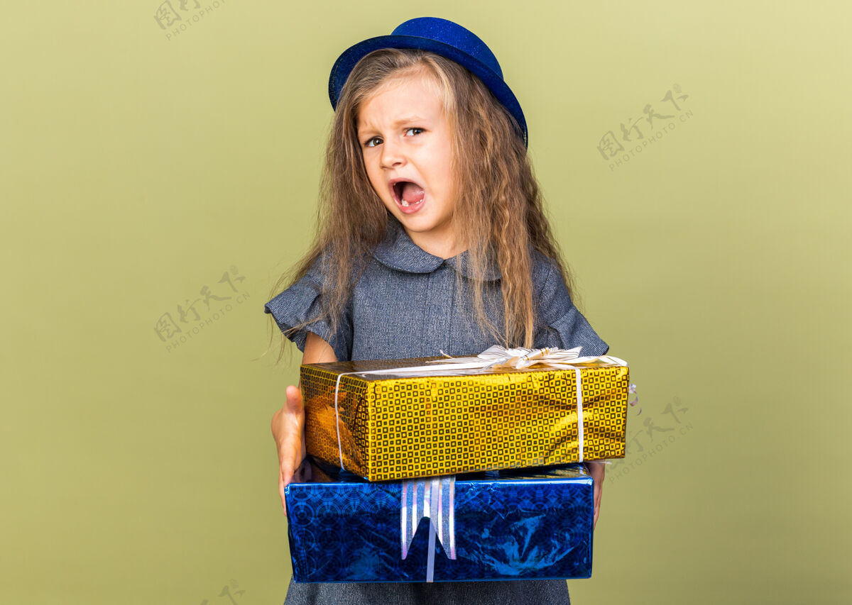 孤立恼怒的金发小女孩 戴着蓝色派对帽 拿着礼品盒 隔离在橄榄绿的墙上 还有复印空间盒子帽子小