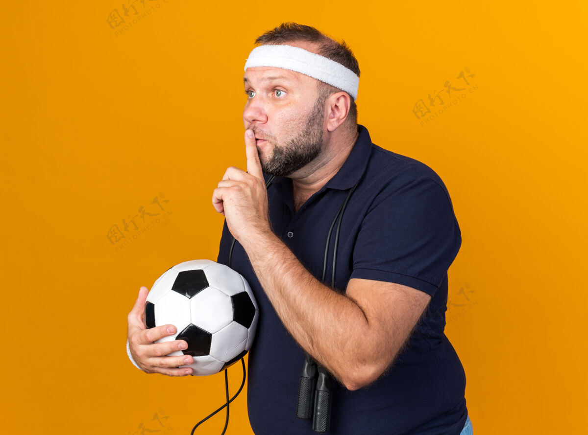 保持被吓坏了的成年斯拉夫运动型男子 脖子上系着跳绳 戴着头带和腕带 拿着球 做着沉默的手势 隔离在橙色的墙上 留着复制空间戴绳子手势
