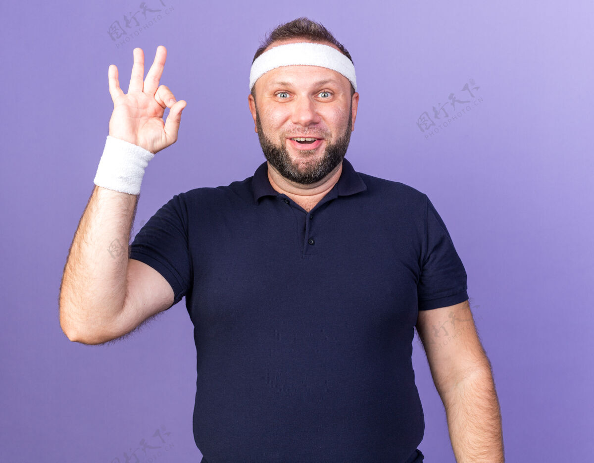 腕带惊讶的成年斯拉夫运动男子戴着头带和腕带表示同意标志隔离在紫色墙上复制空间男人成人穿