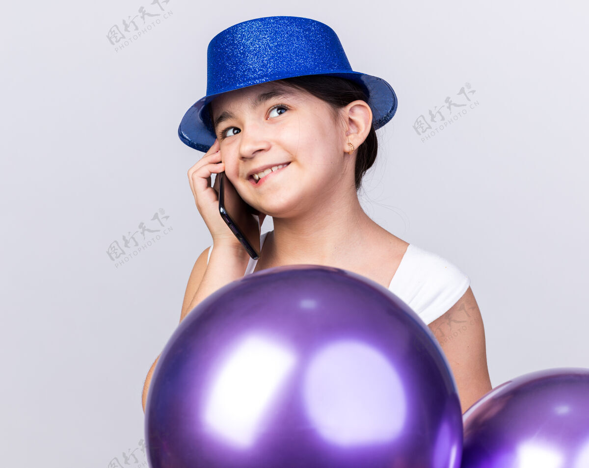 穿高兴的年轻白人女孩戴着蓝色的聚会帽站在氦气球上讲电话隔离在白色的墙壁与复制空间白种人氦孤立