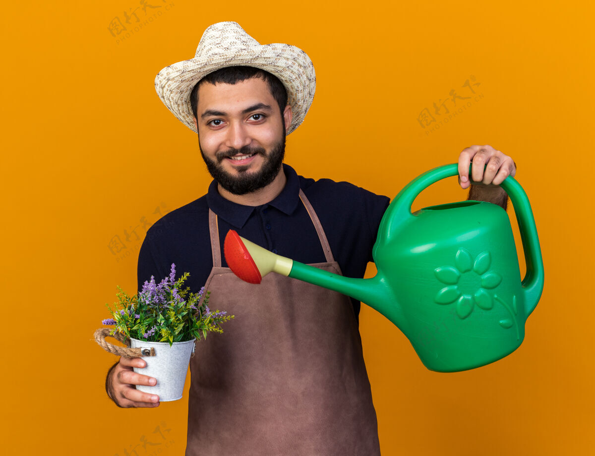 浇水微笑着的年轻白人男性园丁戴着园艺帽在花盆里浇花 可以在橘黄色的墙上独立地复制空间孤立园艺花盆