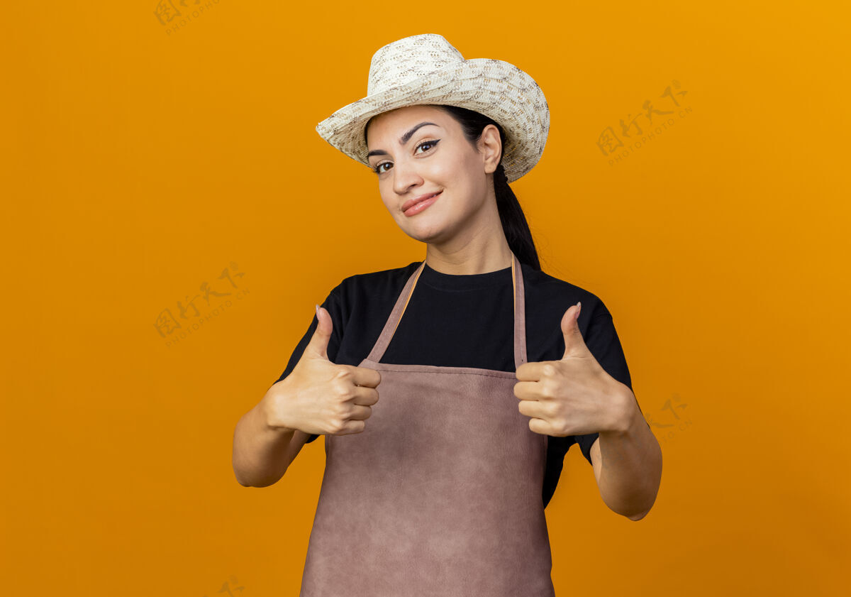 脸年轻漂亮的女园丁围着围裙 戴着帽子 微笑着站在橙色的墙上 竖起大拇指人漂亮女人