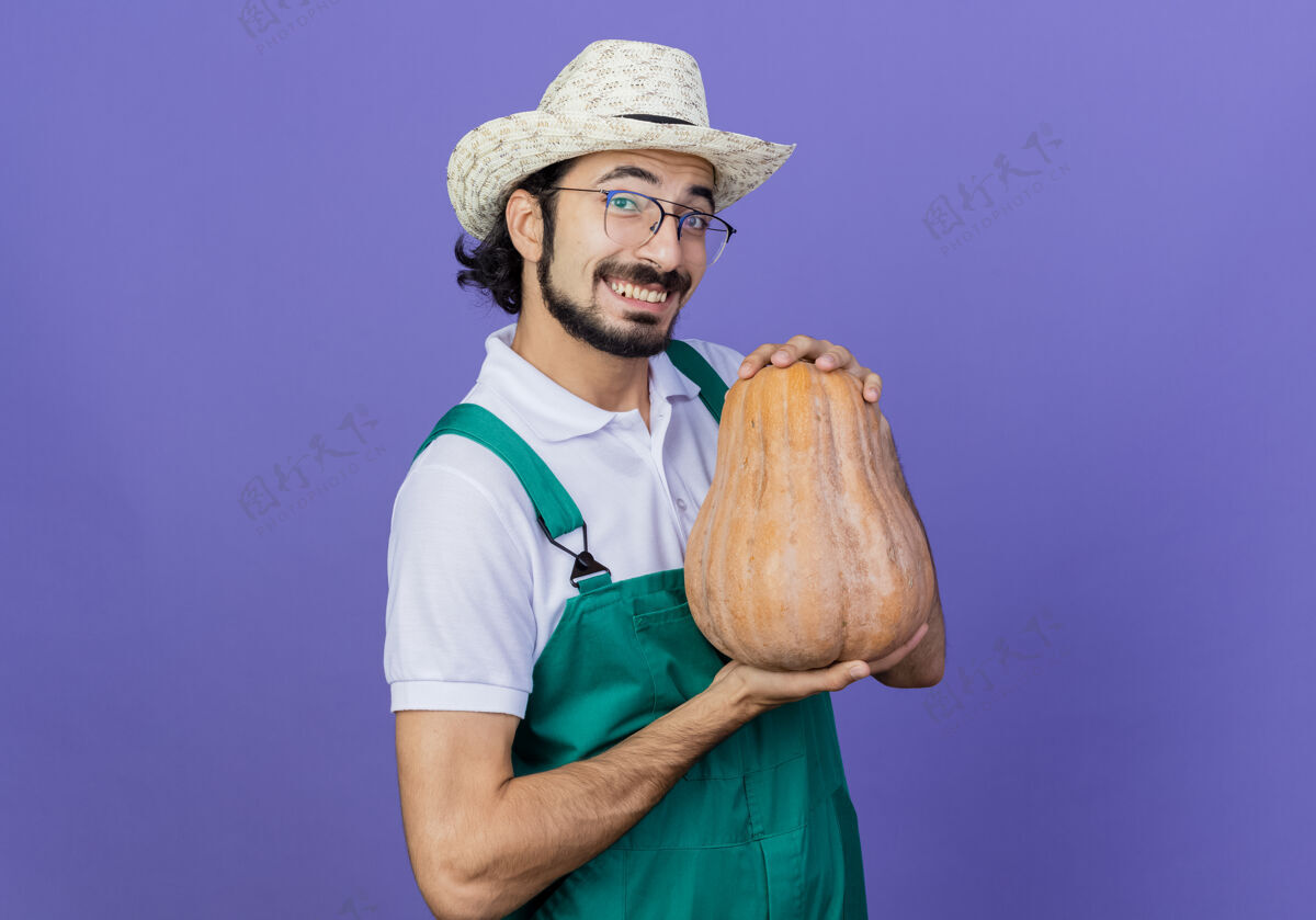 人年轻的留着胡子的园丁 穿着连身衣 戴着帽子 手里拿着南瓜 站在蓝色的墙上 面带微笑地看着前面人胡须抱着