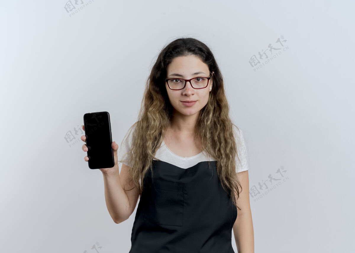 衣服穿着围裙的年轻专业美发师展示智能手机看着前面微笑自信地站在白色的墙上公民女人姿势