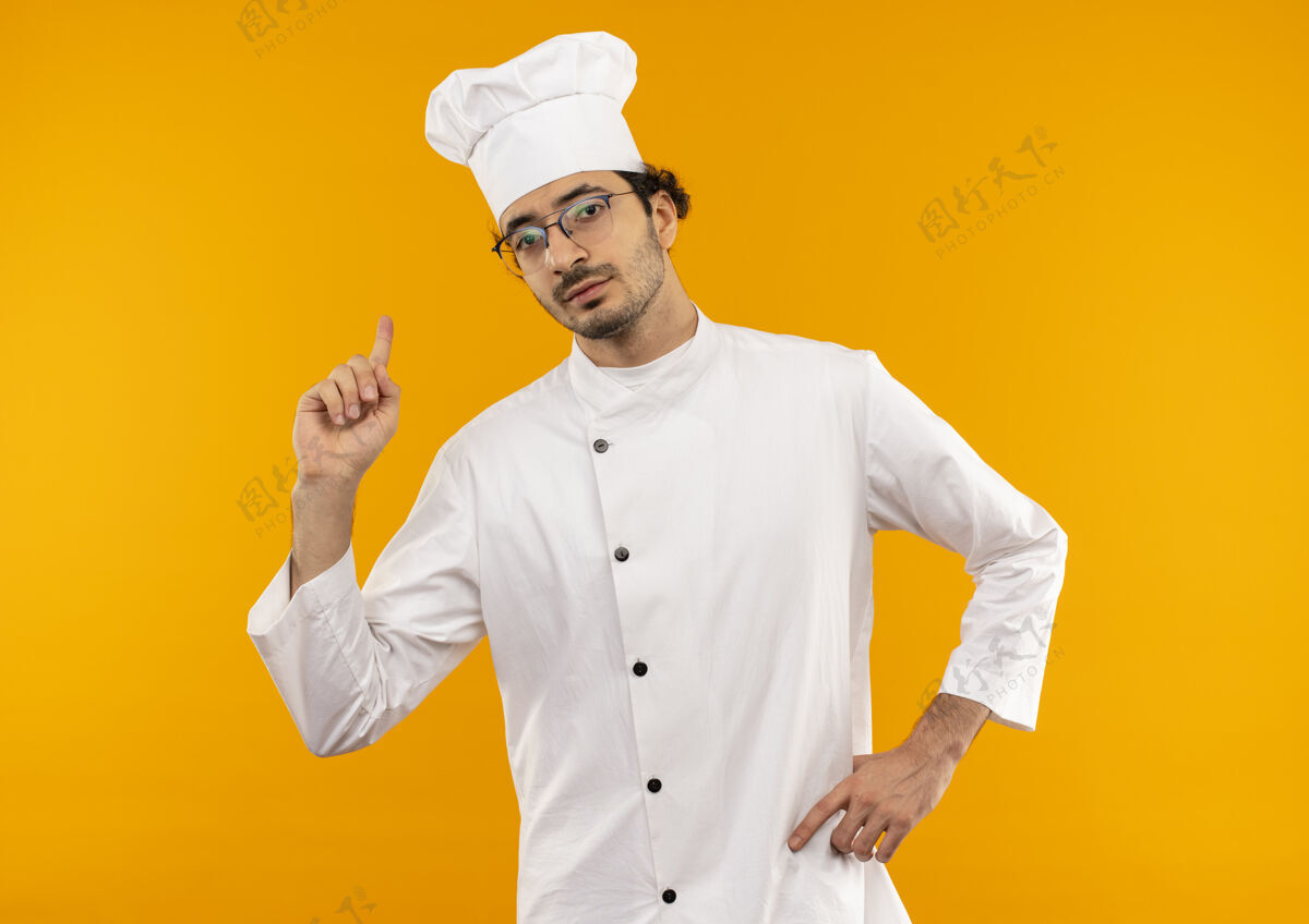 制服自信的年轻男厨师穿着厨师制服 戴着眼镜 指向上方 把手放在臀部自信点厨师