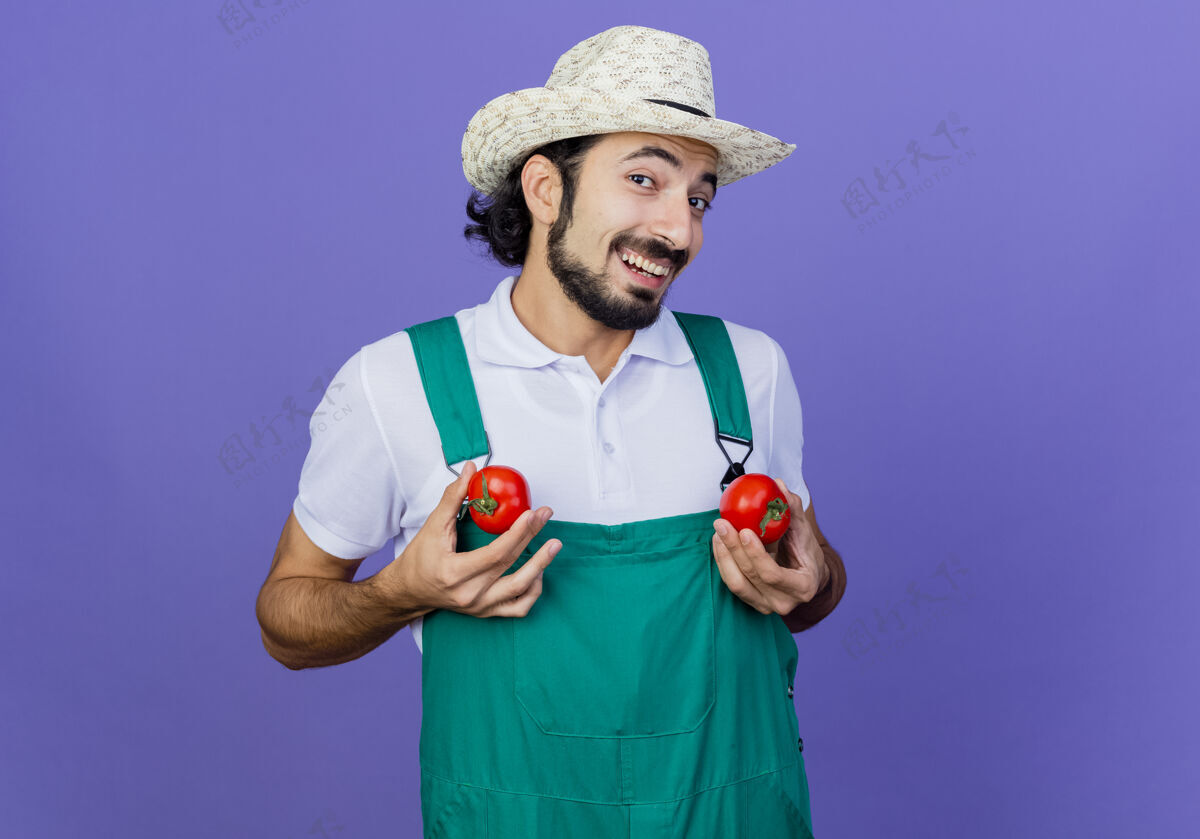 站着有趣的年轻留着胡子的园丁 穿着连体衣 戴着帽子 手里拿着新鲜的西红柿 站在蓝色的墙上 兴高采烈地微笑着看着前面人花园感觉