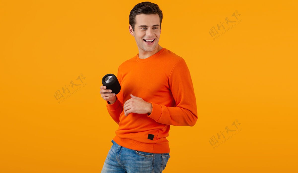 科技穿着橙色毛衣的时髦微笑的年轻人拿着无线扬声器快乐地听音乐享受橙色的乐趣搞笑乐趣扬声器