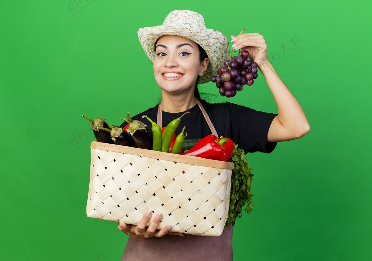 人年轻漂亮的女园丁围着围裙 戴着帽子 抱着装满蔬菜和葡萄的篮子快乐而积极地站在绿色的墙上市民帽子举行
