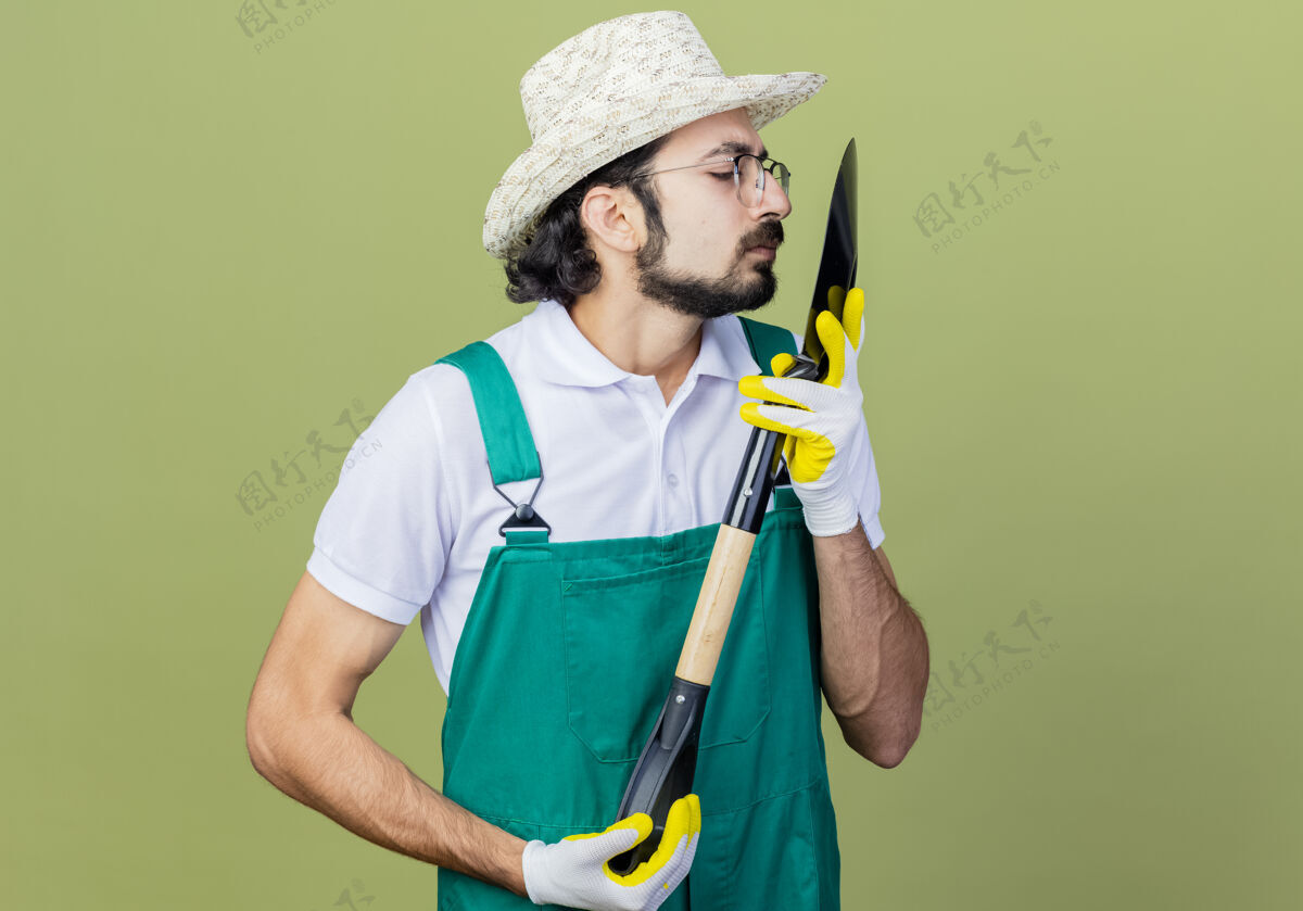 光年轻的留着胡须的园丁穿着连体衣 戴着帽子 手里拿着铲子 站在淡绿色的墙上近距离地看着它封闭人市民