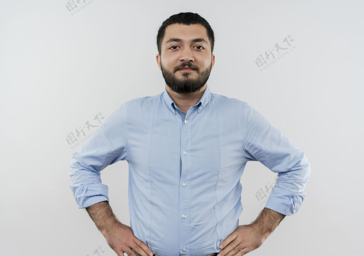 站着身穿蓝色衬衫 留着胡须的年轻人站在白色的墙上 面带微笑 自信地看着前面 双臂放在臀部人男人衬衫