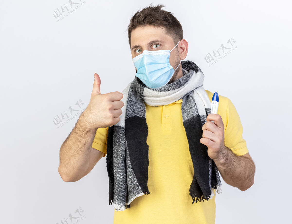 举行年轻的金发病患戴着医用面罩和围巾 竖起大拇指 拿着隔离在白墙上的温度计疾病体温计围巾
