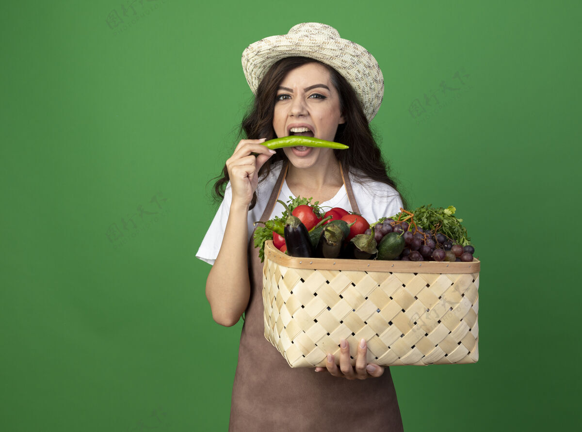 人穿着制服的年轻漂亮的女园丁戴着园艺帽 手里拿着菜篮子 假装咬隔离在绿墙上的辣椒花园咬园艺